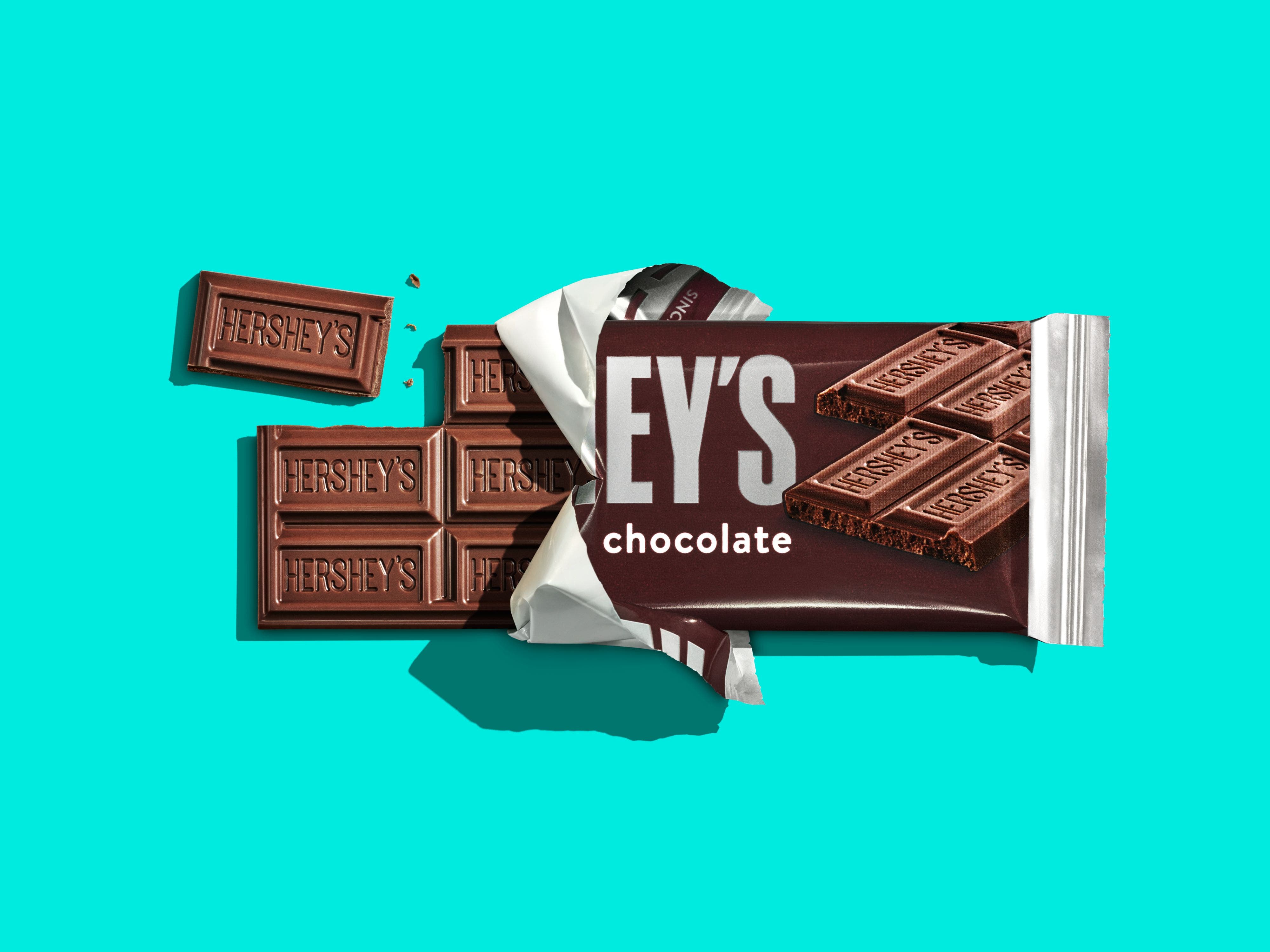 Barra de chocolate HERSHEY’S Ao Leite, com embalagem na metade e um pedaço solto.